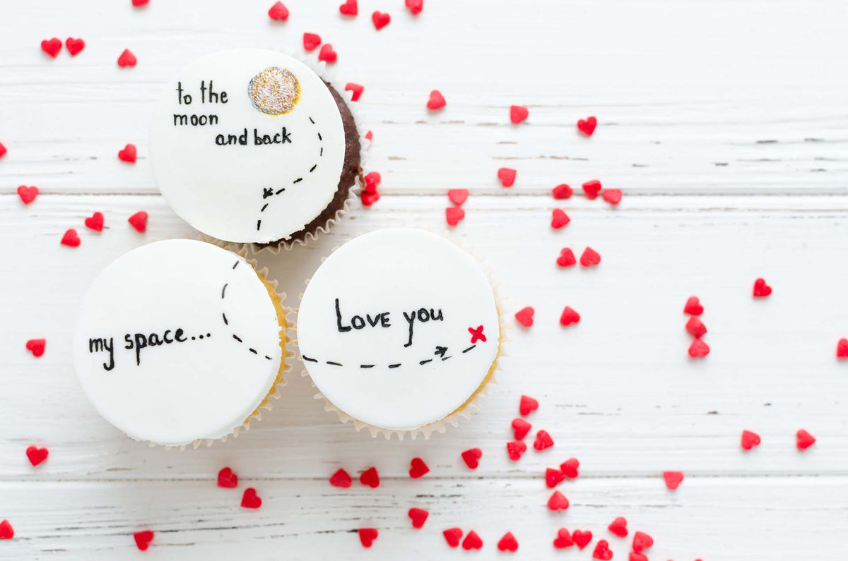 Drei Cupcakes mit bedruckten Esspapier-Auflegern und roten Herzstreuseln auf weißem Holz