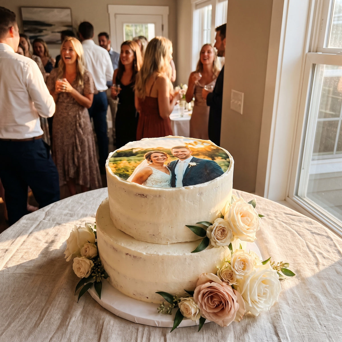 Zweistöckige Hochzeitstorte mit rundem essbaren Paarfoto-Aufleger und Rosen, Feier im Hintergrund